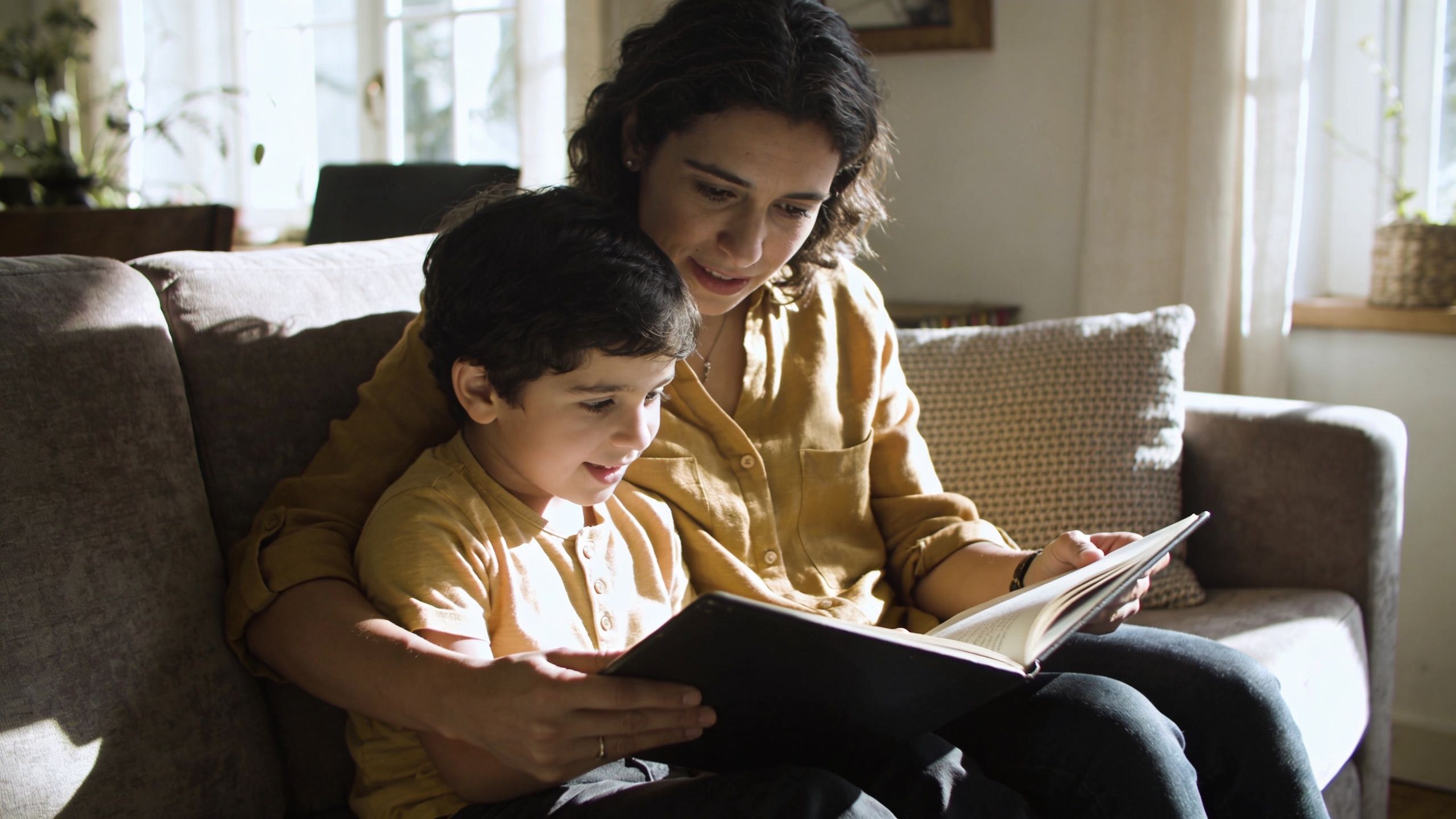 A mother sitting on a couch with her young son, reading a book together in a sunlit room.