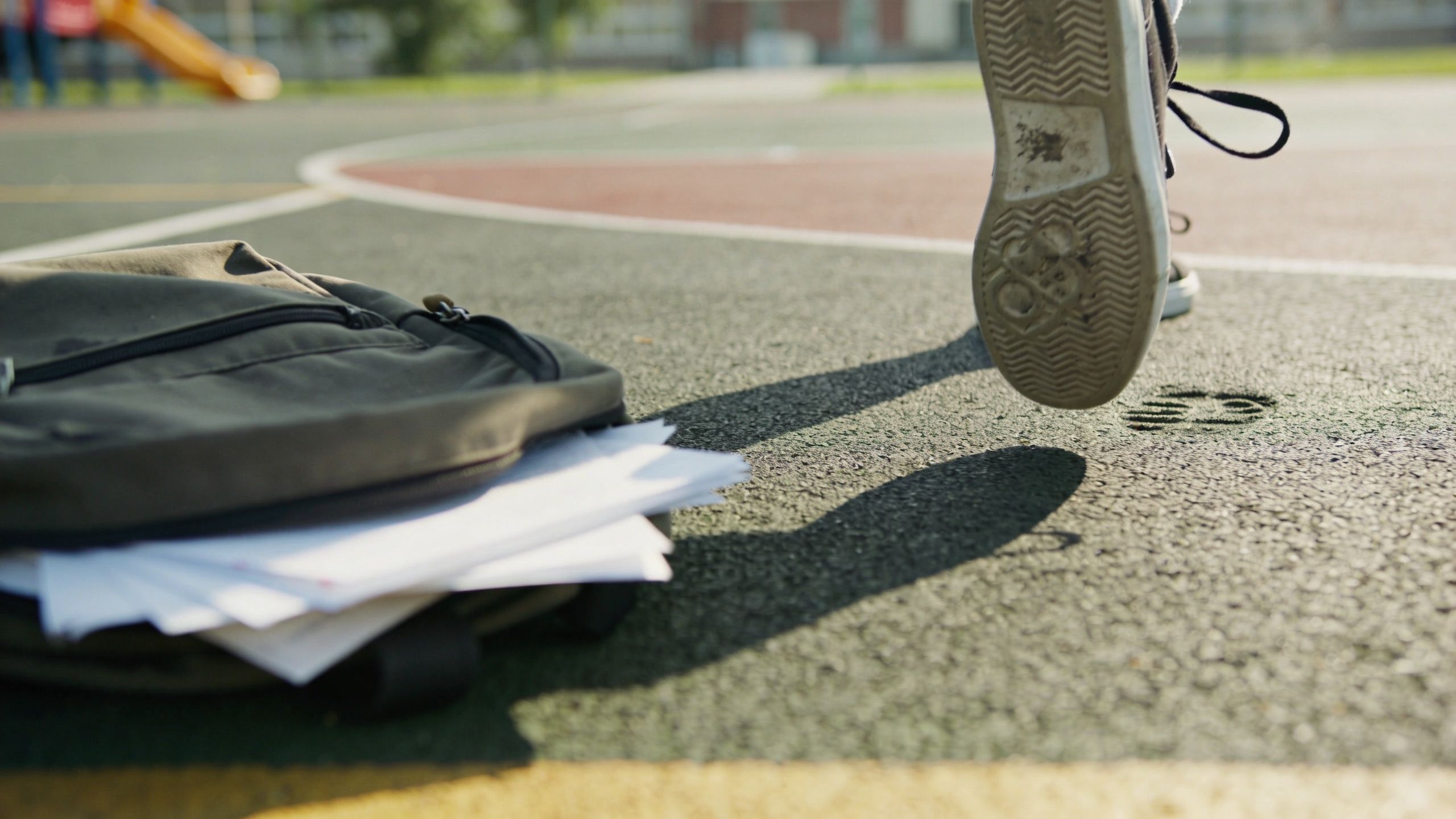 A student's backpack with loose papers left on a playground while someone walks away in the distance.