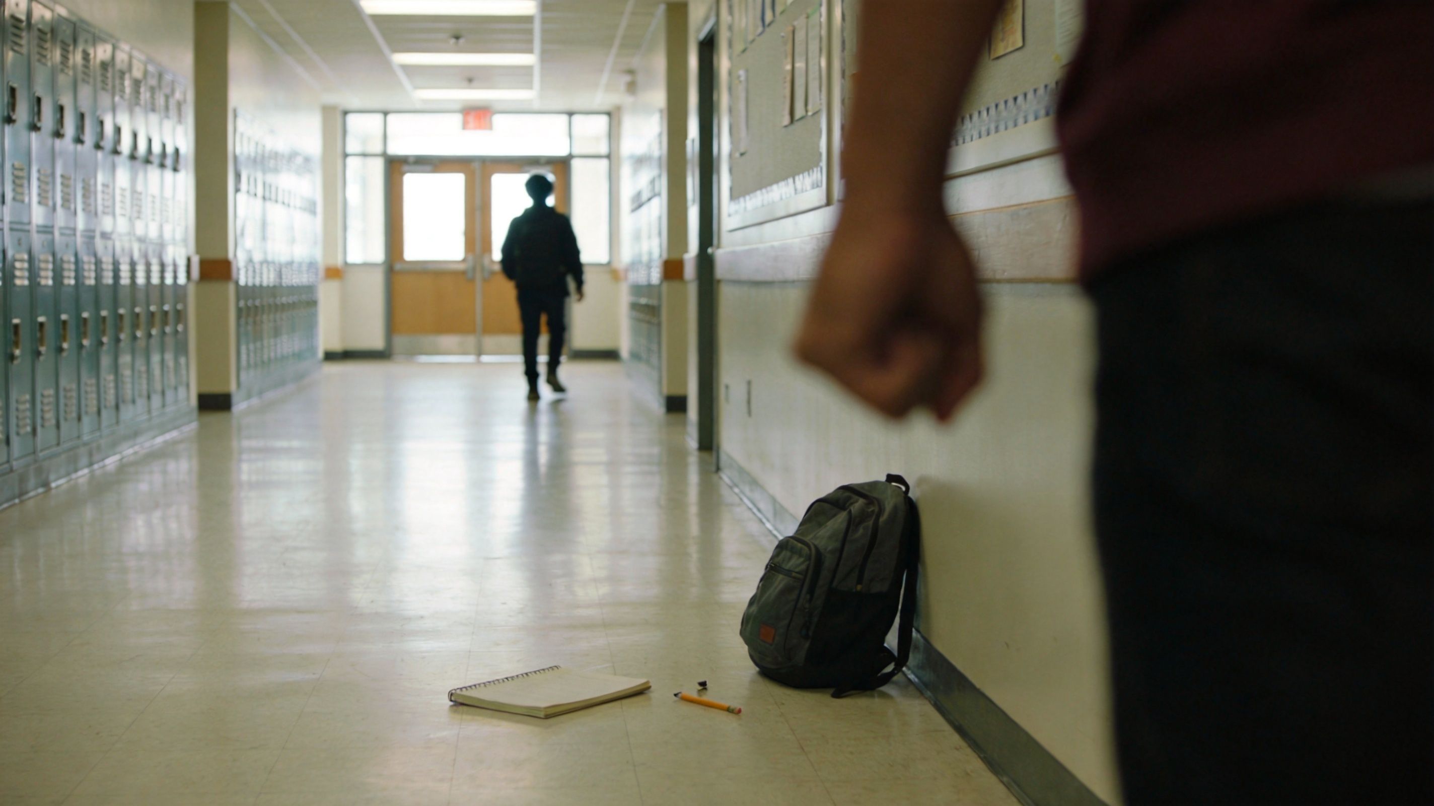 A close-up view of a clenched fist with a student's abandoned backpack and notebook in a school hallway.