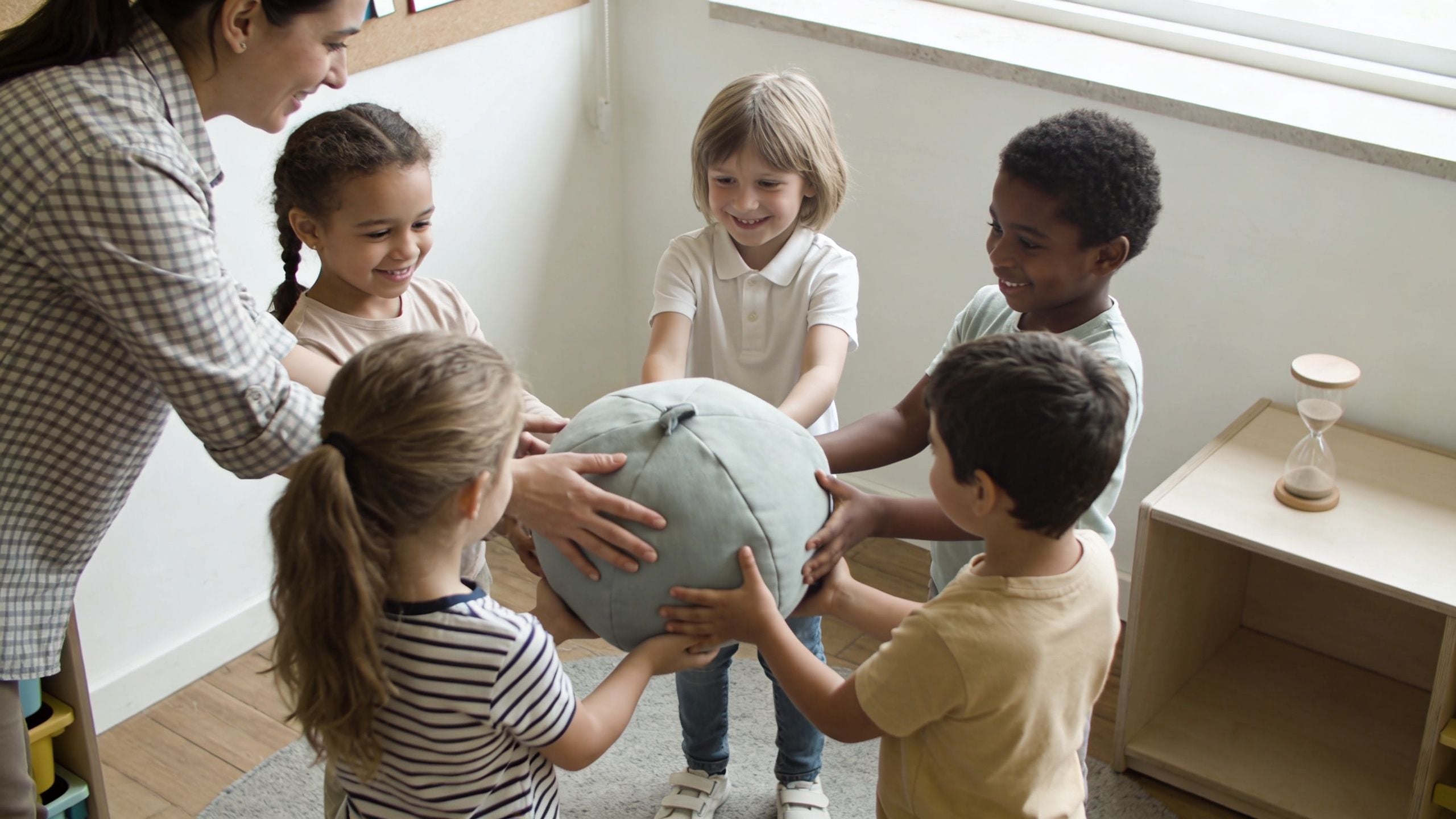 A teacher and a group of young preschoolers playing a collaborative game with a soft grey ball.