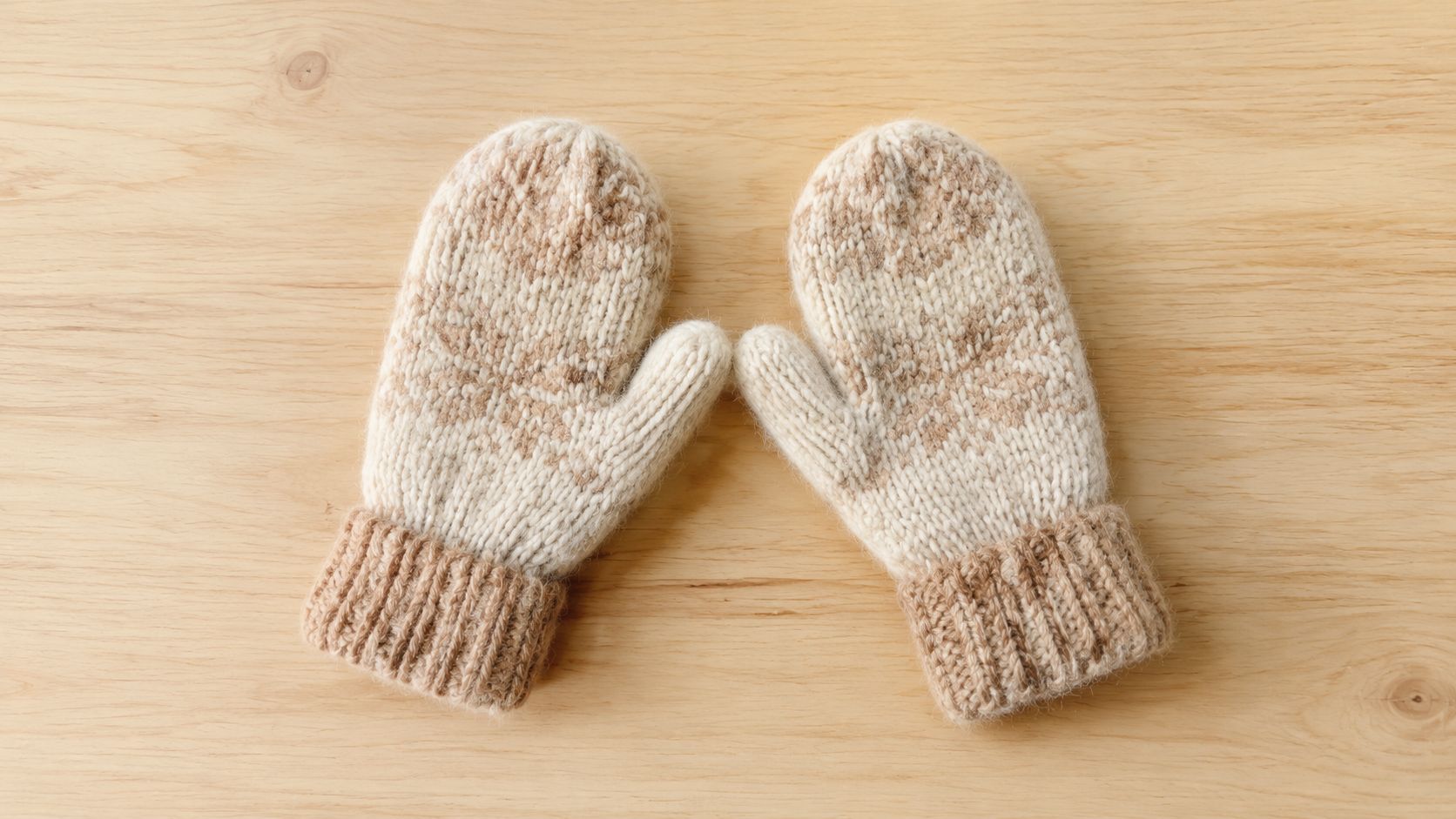 A pair of cozy, cream and light brown knit winter mittens resting on a light wooden background.