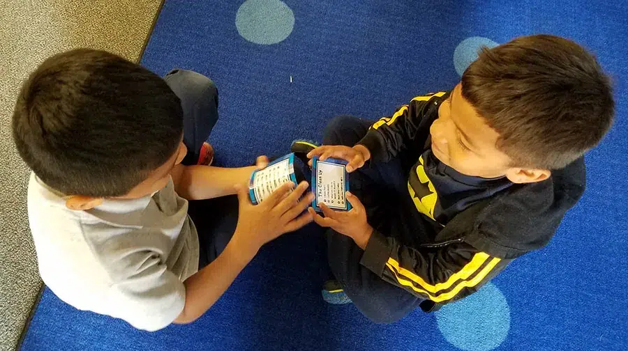 Two students sit facing each other on a classroom carpet, each holding a Soul Shoppe Clean Up card as they work through a conflict.